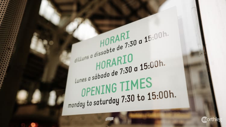 Multilingual opening hours sign in green and black text, displaying times from 7:30 to 15:00 for Monday to Saturday.