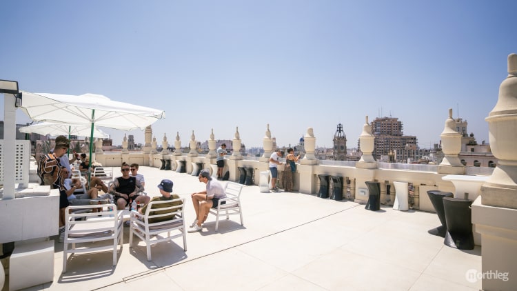 Rooftop terrace with white chairs, umbrellas, and decorative stone elements, overlooking Valencia's urban landscape on a bright, clear day.