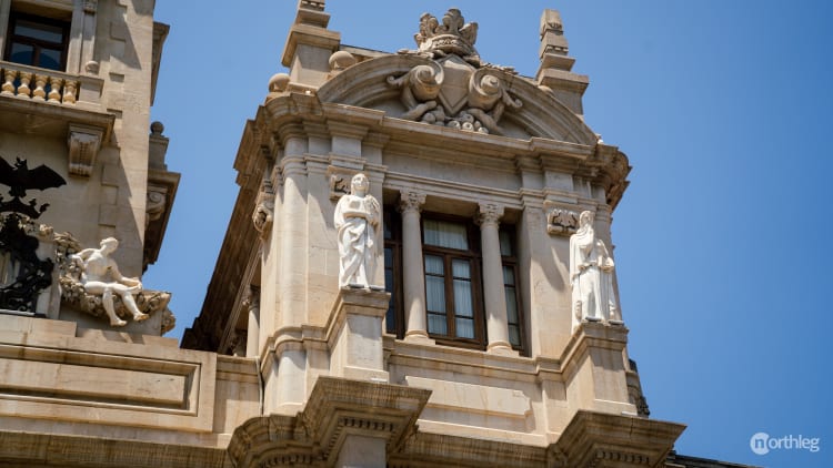 Baroque-style building facade with white stone statues, decorative architectural elements, and classical columns against a bright blue sky.