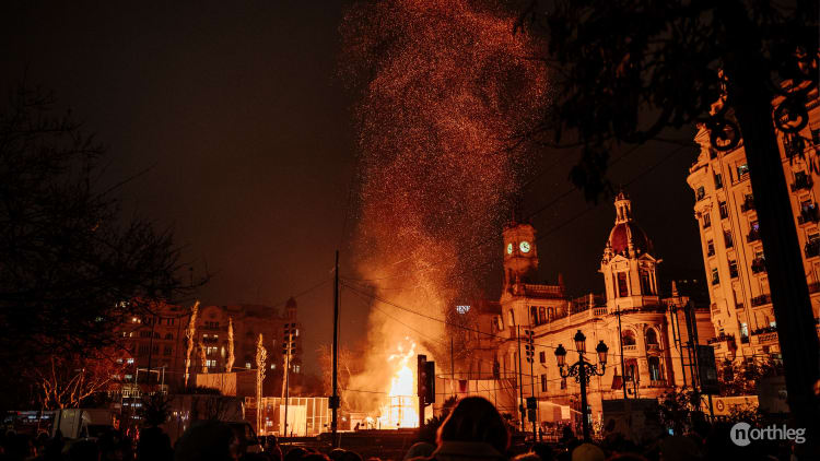 Nighttime image of a large fire burning in Valencia's Plaza del Ayuntamiento during the Fallas festival, with historic buildings and a shower of burning embers in the background.