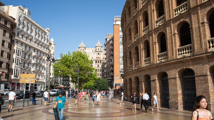 Brick bullring building in Valencia with ornate arched windows, surrounded by historic city streets and pedestrians on a sunny day.