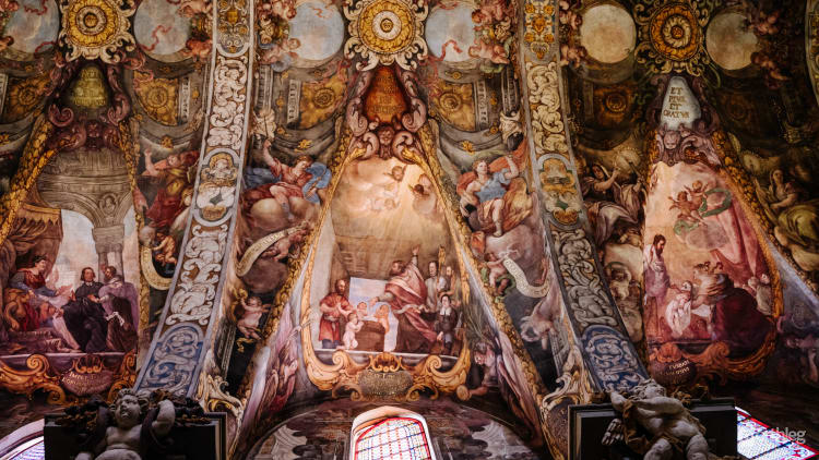 Elaborate baroque ceiling fresco with ornate religious paintings, featuring multiple scenes of saints and biblical narratives in warm golden and pastel tones.