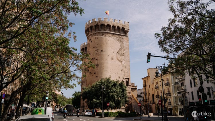 Large stone cylindrical medieval tower with crenellated top, situated in an urban street setting with trees and traffic signals.