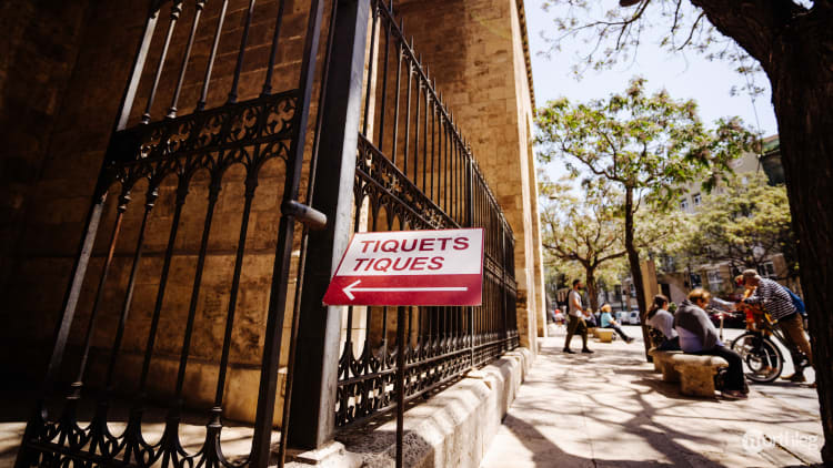 Red ticket sign pointing to entrance of Torres de Serranos with ornate iron fence and stone walls in sunlight