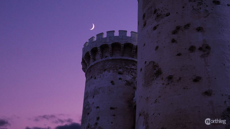 Medieval stone towers with crenellated tops against a purple twilight sky with a crescent moon, showing historic city fortifications.