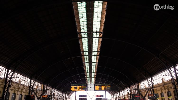 Interior view of a grand train station with a high arched glass and metal roof, featuring information boards and a central clock.