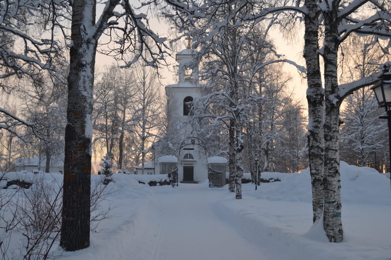 En vinterbild från en kyrkogård med ett kyrktorn i bakgrunden. Ett snötäckt spår leder fram till en port som är en del av ett staket runt kyrkan och kyrkogården. Träd med snö på grenarna ramar in bilden. Mark och ytor är täckta av snö. Himlen i bakgr