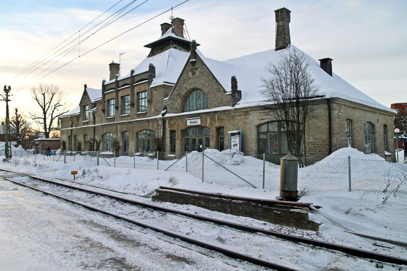 En bild visar ett snötäckt landskap med en stor byggnad i bakgrunden. Byggnaden är gjord av ljus tegel eller sten med ett brant tak täckt av snö. Det finns flera skorstenar synliga på taket. Arkitekturen inkluderar stora, välvda fönster och en ingång