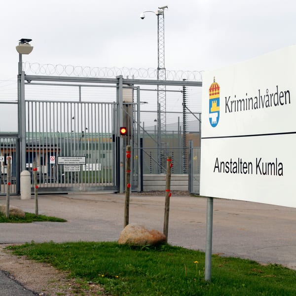 En bild visar entrén till en institution med en stor, vit skylt till höger som texten "Kriminalvården" och "Anstalten Kumla" samt Kriminalvårdens logotyp. Bakom skylten och till vänster finns ett högt stängsel med rakbladstråd överst och en grind som