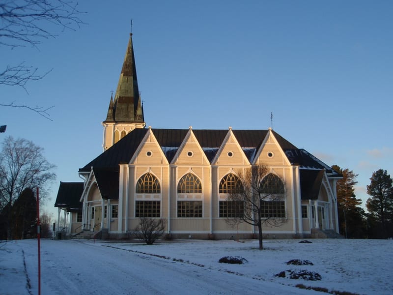 Bilden visar en gulbeige kyrka med mörkt tak och en spetsig kyrkspira, sedd från sidan. Marken runt kyrkan är täckt av snö, och en snötäckt grusväg leder fram till byggnaden. Det är soligt och himlen är klarblå. Träd med kala grenar står i förgrunden