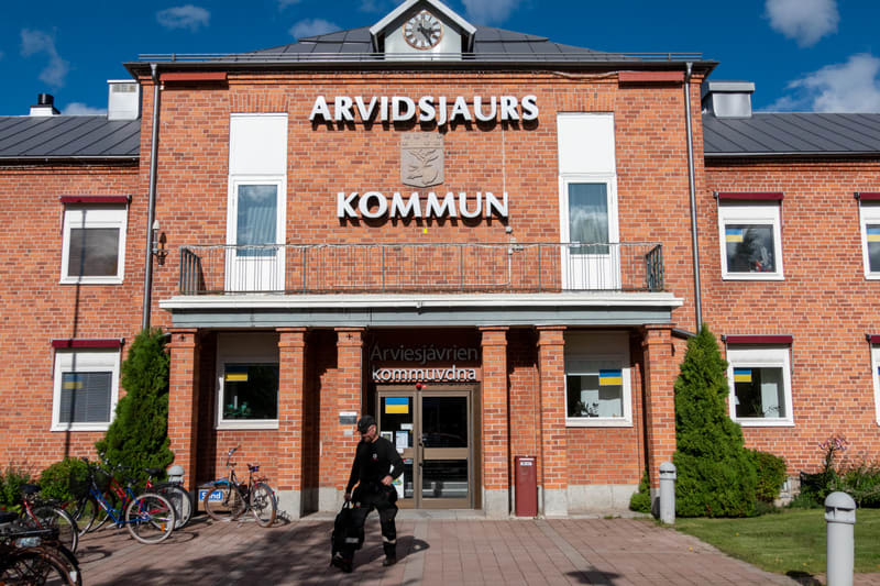 En fasadbild av en större röd tegelbyggnad med texten "ARVIDSJÄURS KOMMUN" i stora vita bokstäver ovanför entrén. Mellan orden finns en grå vapensköld. Ovanför texten sitter en klocka. Entrén består av två glasdörrar, och över denna finns en balkong