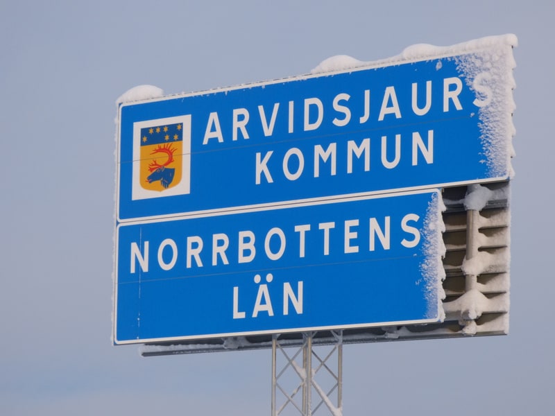 Bilden visar en blå vägskylt monterad på en metallstolpe. Skylten har vit text som lyder "ARVIDSJAUR KOMMUN" och "NORRBOTTENS LÄN". Skylten har också en vapensköld som föreställer en röd renhornskrona omgiven av gula stjärnor på en gul bakgrund, samt