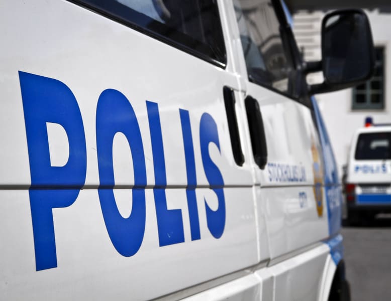 Bilden visar ett närperspektiv på sidan av en vit polisbil med blå text som stavar "POLIS" i förgrunden. En del av texten 'STOCKHOLMS LÄN' är synlig under 'POLIS'. Bilens fönster och dörrhandtag är också synliga. I bakgrunden, suddigt, syns en annan