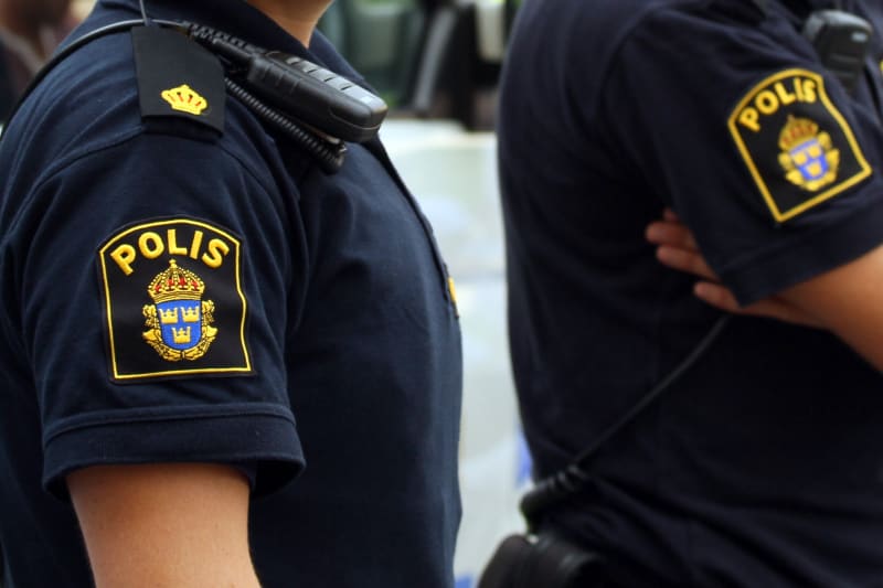 Bilden visar närbilder på två individer iklädda mörkblå pikétröjor som ser ut att vara en del av polisuniformen. På vänster arm finns en gul och blå vapensköld med texten 'POLIS' ovanför, vilken är Sveriges polismyndighets emblem. Ovanför emblemet, p