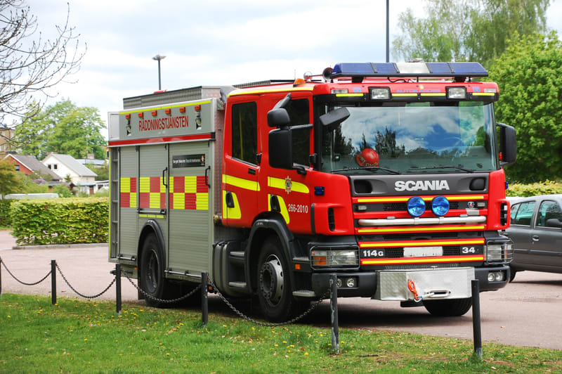 Bilden visar en röd brandbil av märket Scania, med texten "RÄDDNINGSTJÄNSTEN" tydligt skriven på sidan i rött med silverfärgad bakgrund. Fordonet har även gula och röda rutmönster på sidan samt gula reflexband. På fronten syns två blåa extraljus och