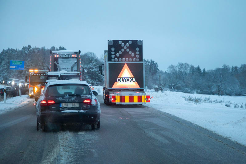 Bilden visar en snötäckt väg i vinterlandskap. I förgrunden syns bakdelen av en mörk personbil, täckt med snö, körande på en snöig och isig vägbana. Framför personbilen står en lastbil med en stor varningsskylt som visar texten "OLYCKA" inuti en röd
