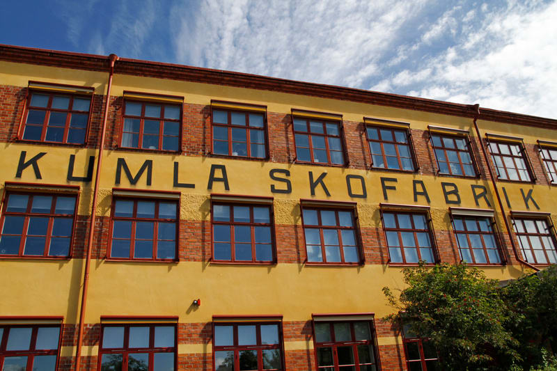 Bilden visar fasaden på en gul byggnad med tegelinslag och många fönster. På den gula fasaden står texten "KUMLA SKOFABRIK" i stora svarta bokstäver. Byggnaden har flera rader med fönster med mörka ramar, och vissa fönster har utvändiga persienner el