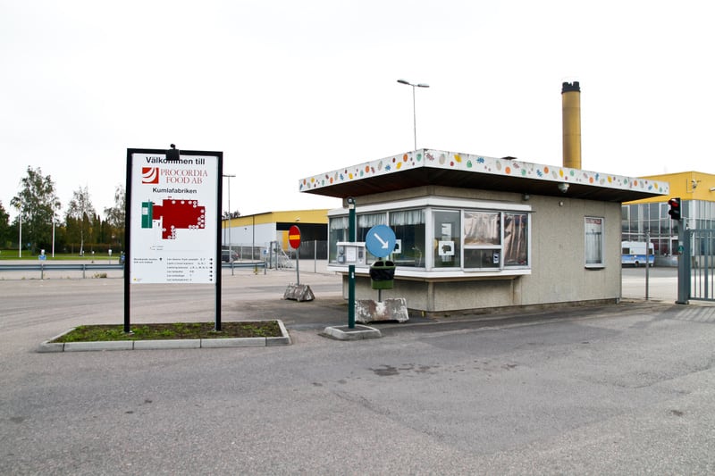 Bilden visar ett fabriksområde med en skylt som välkomnar till Procordia Food AB Kumlafabriken. En grindstuga eller vaktkur står till höger om skylten, med fönster och en dekorerad takfot. Utanför vaktkuren finns en stolpe med en blå pil som pekar ne
