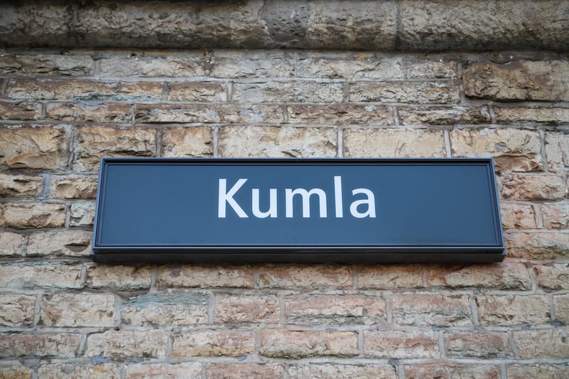 Bilden visar en rektangulär skylt med texten "Kumla" i vit färg på en mörkgrå bakgrund. Skylten är monterad på en tegelvägg som består av oregelbundna, ljusbruna stenar. Stenarna varierar i storlek och form, och fogarna mellan dem är synliga. Ljuset