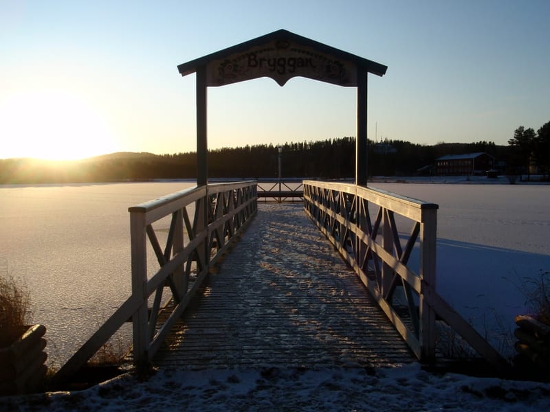 Bilden visar en träbrygga som sträcker sig ut över en frusen sjö. Bryggan har sidoräcken och en takkonstruktion över ingången med texten "Bryggan" och dekorativa inslag. Viss snö och is ligger på bryggans plattform. Solen, som står lågt på himlen, ly
