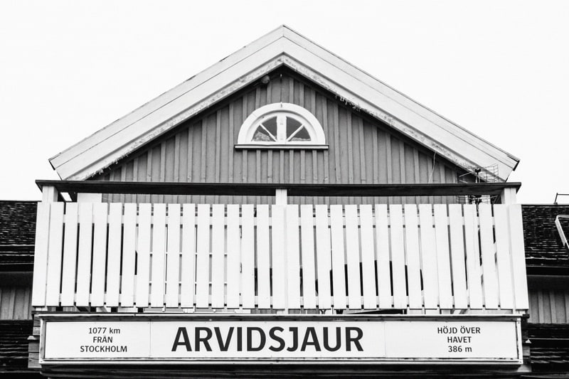 Bilden är tagen i svartvitt och visar den övre delen av en byggnad med en takgavel och en balkong. På takgaveln sitter ett halvcirkelformat fönster med spröjs. Under fönstret finns en träpanel i lodräta plankor. En vit träbalkong sträcker sig horison