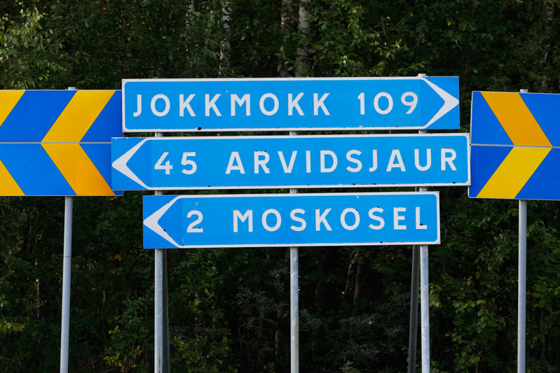 Bilden visar tre blåa vägskyltar med vit text och pilar som är monterade på metallstolpar. Den översta skylten anger "JOKKMOKK 109" med en pil som pekar åt höger. Mellan skylten visar "45 ARVIDSJAUR" med en pil som pekar åt vänster. Den understa skyl
