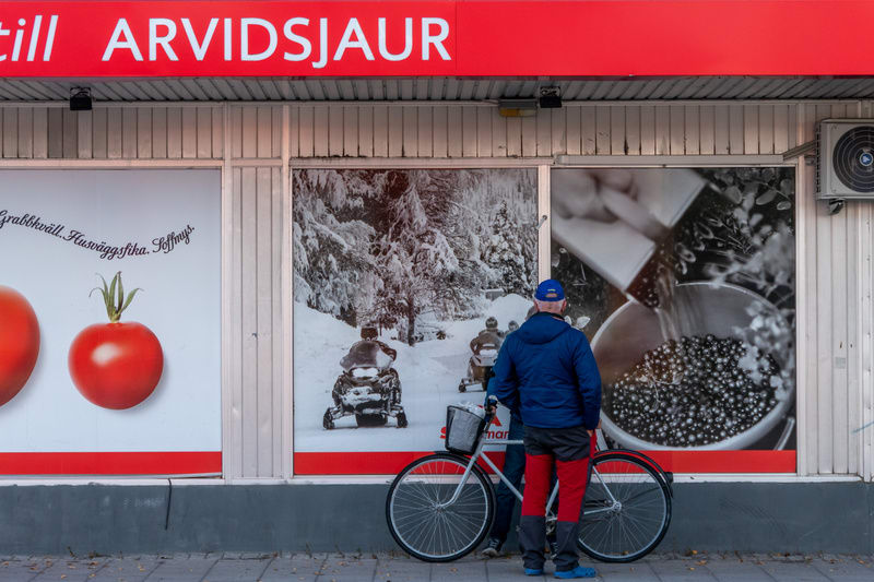 En person, sedd bakifrån, står på en trottoar framför en byggnad med butiksfönster. Personen bär en blå jacka, röda byxor, blå skoskydd och en blå keps. En cykel med främre korg står parkerad bredvid personen. Ovanför butiksfönstren sitter en röd sky