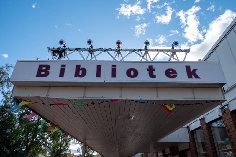 Bilden visar fasaden på en byggnad med en skylt där ordet 'Bibliotek' står skrivet i mörkröda bokstäver på en vit bakgrund. Ovanför skylten finns en metallställning med flera runda lampor som har blå eller röd centrum. Under skylten, fäst vid en utsk