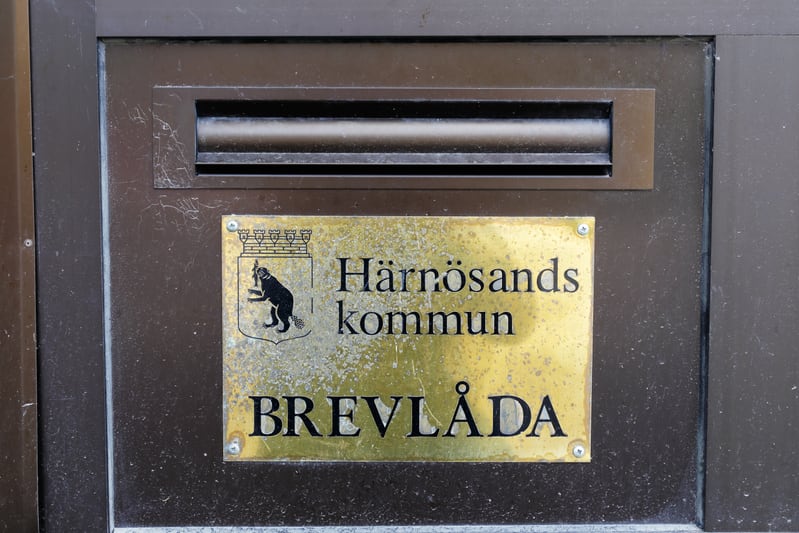 Bilden visar en detalj av en mörkbrun dörr eller väggyta med en guld- eller mässingsfärgad skylt fäst med fyra skruvar. Ovanför skylten sitter ett horisontellt, rektangulärt brevinkast. Skylten är smutsig och har texten "Härnösands kommun BREVLÅDA".