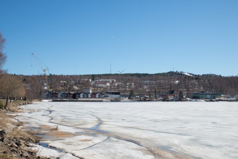 Bilden visar ett landskap med en delvis isbelagd sjö eller ett vattendrag i förgrunden, som sträcker sig från vänster till förgrunden i mitten. Marken till vänster om vattnet är gräsbevuxen med nakna träd och buskar, samt stenar längs strandlinjen. I