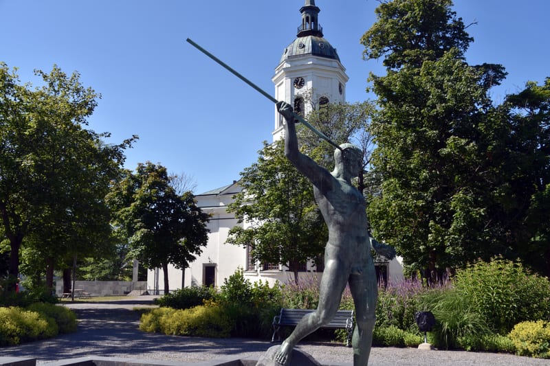 Bilden visar en bronsstaty av en man med ett spjut i förgrunden, till höger i bilden. Staten står på en stenbas. I bakgrunden syns en vit kyrkobyggnad med ett klocktorn och en kupol med ett kors. Grönskande träd omger kyrkan och statyn, och marken be