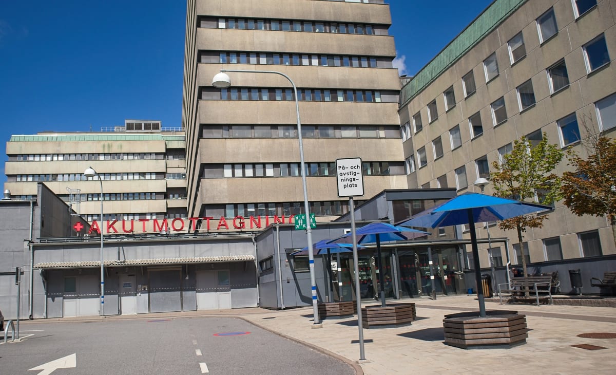 Bilden visar entréområdet till en byggnad som verkar vara en akutmottagning, framför en större sjukhusliknande byggnad under klarblå himmel. Ett rött skyltfönster med texten 'AKUTMOTTAGNING' och ett rött kors är placerat över en entré. Ett trafikmärk