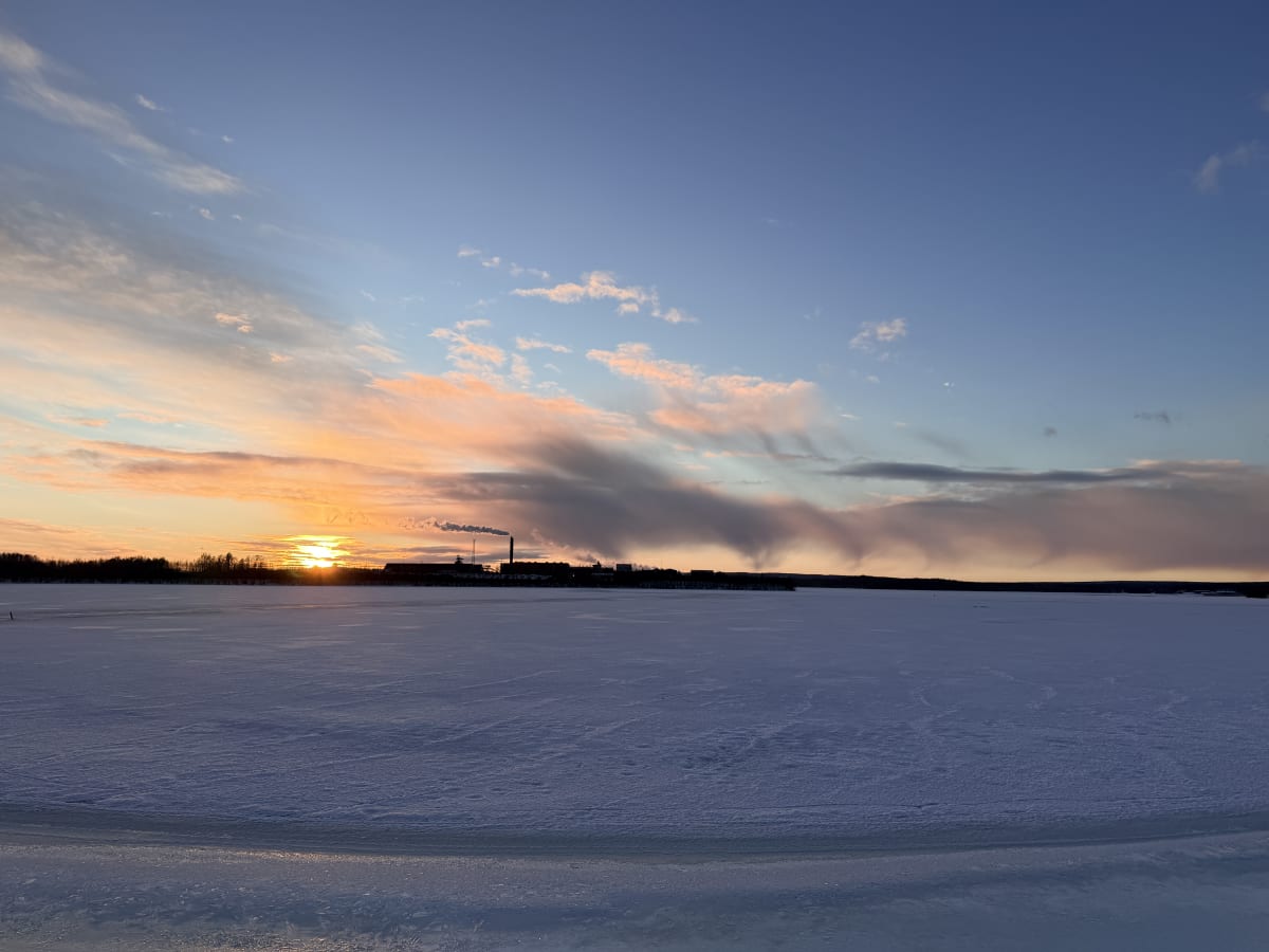 Bilden visar ett vinterlandskap med en stor, snö- och isbetäckt yta i förgrunden som kan vara en sjö eller ett vattendrag. Horisonten domineras av en mörk silhuett av träd och vad som ser ut som byggnader, varav en del avger rök eller ånga från en sk