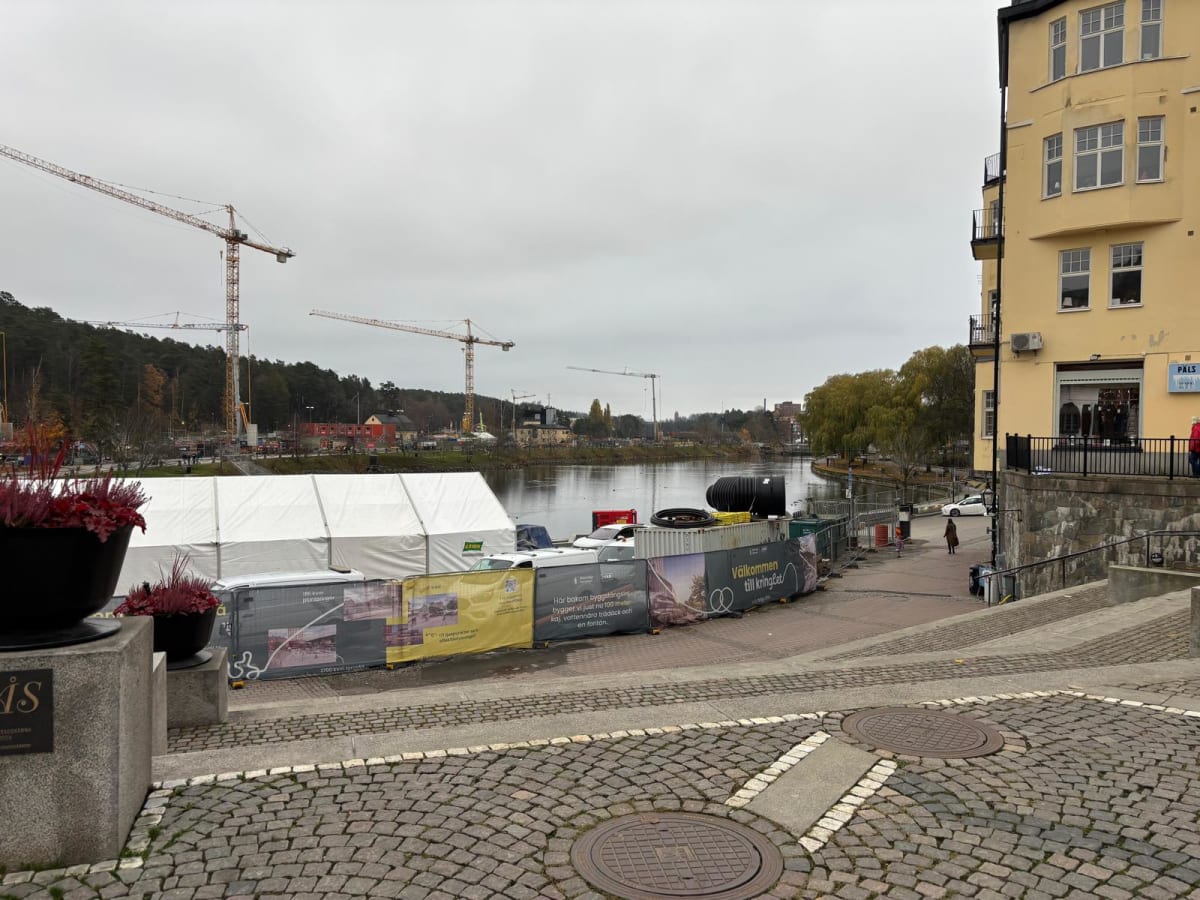Bilden visar ett stadslandskap med vatten, byggnader och byggarbetsplatser under en molnig himmel. I förgrunden syns en stenbelagd yta med brunnslock och planteringar med rödaktiga växter. Till höger finns en gul byggnad med flera fönster och balkong