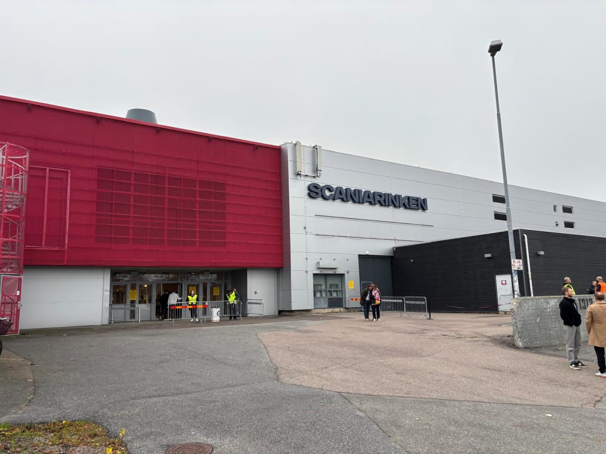 Bilden visar utsidan av en stor byggnad med namnet "SCANIARINKEN" skrivet i stora bokstäver på den grå fasaden. Delar av byggnadens fasad är röd med en perforerad yta och ett utskjutande rött spir altrappliknande element syns till vänster. Framför by