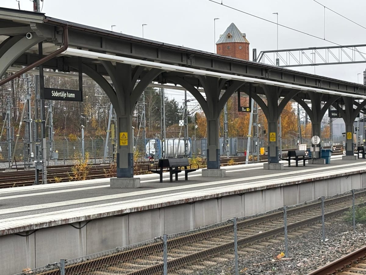 Bilden visar en järnvägsstation från en upphöjd synvinkel. I förgrunden syns tågrälsar avskärmade av ett metallstängsel. Ovanför stängslet, i mitten av bilden, finns en plattform med ett tak konstruerat av grå valv och pelare. Under taket, på plattfo