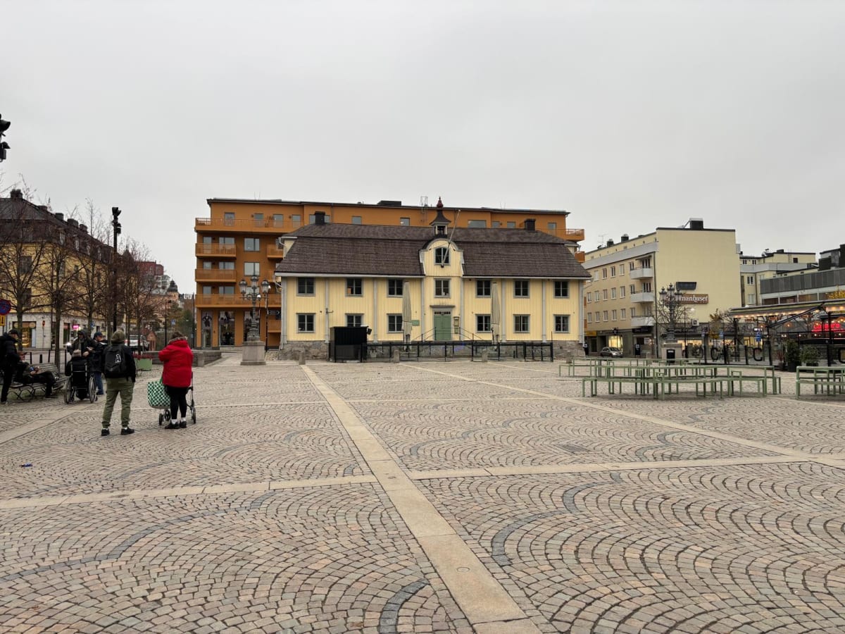 Bilden visar ett torg med gatsten och byggnader i bakgrunden. I mitten av torget står en äldre, gul byggnad med brunt tak och vita fönsterkarmar. Framför denna byggnad syns grindar och vad som verkar vara parasoller hopfällda. Till vänster om den gul