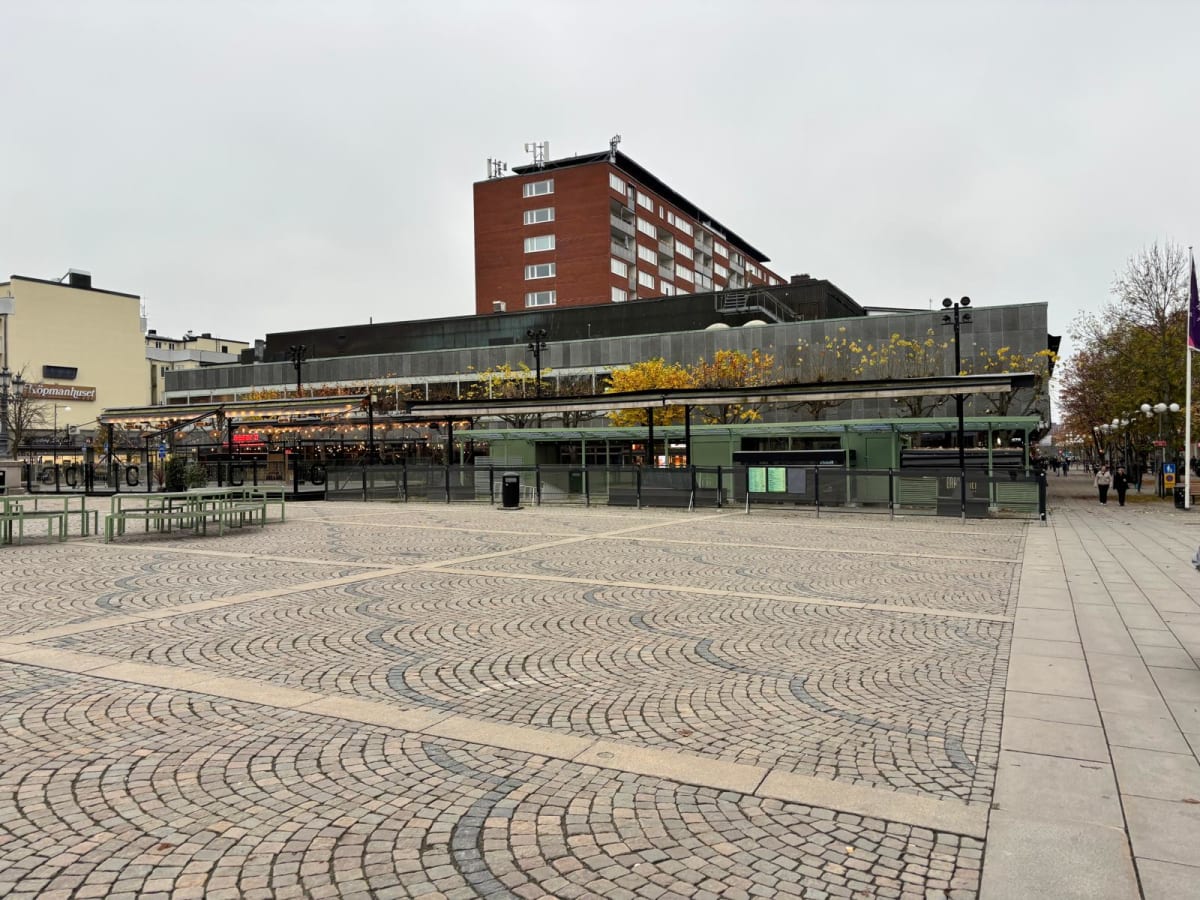 Bilden visar ett torg med gatstenar i ett cirkulärt mönster. I förgrunden till vänster finns flera tomma, ljusgröna metallbord och bänkar. Bakom dessa syns en byggnad som ser ut att vara ett köpcentrum eller liknande kommersiell byggnad, med skyltfön