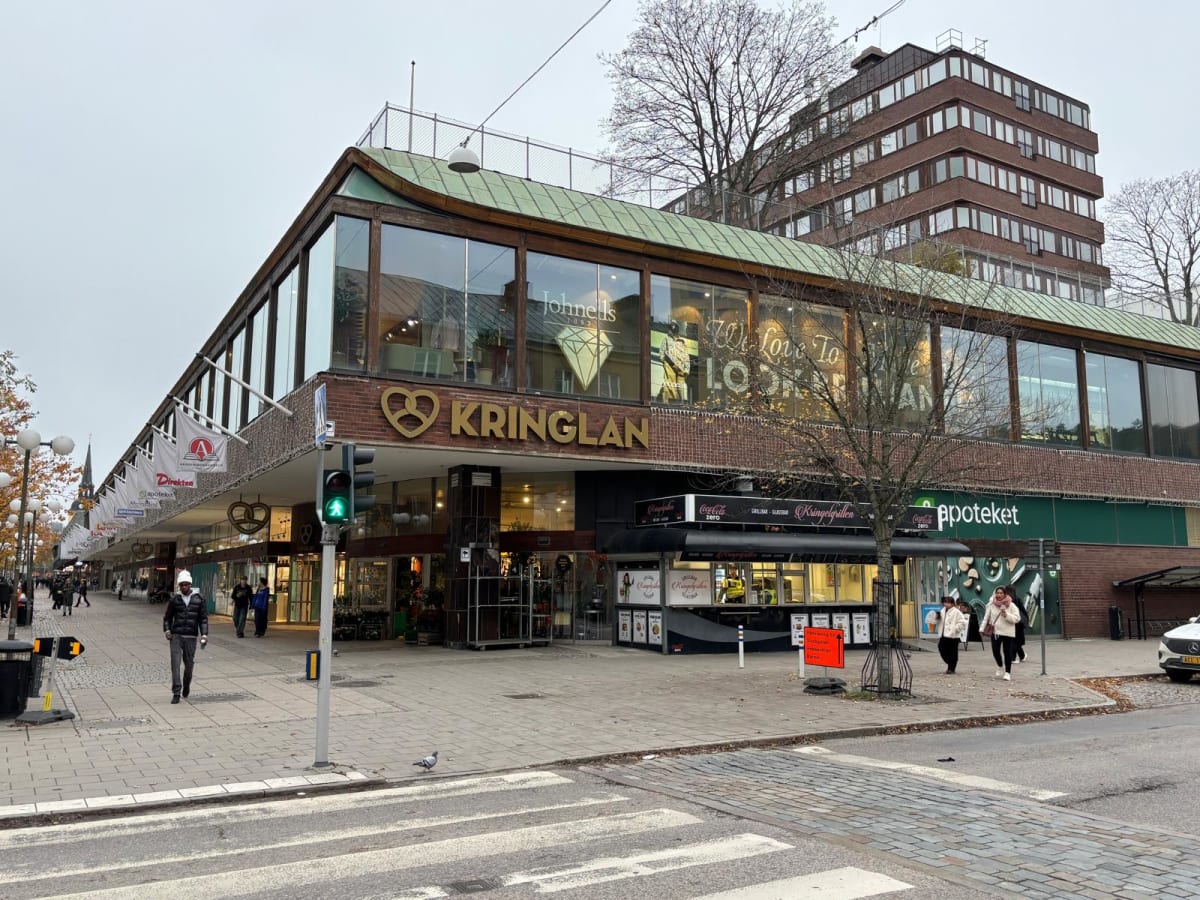 En bild tagen utomhus under dagtid, som visar en gata med en butiksfasad och trottoar. Huvudbyggnaden har utskjuten övervåning med stora fönster och en grön takyta. På fasaden finns texten "KRINGLAN" i stora guldfärgade bokstäver, tillsammans med en