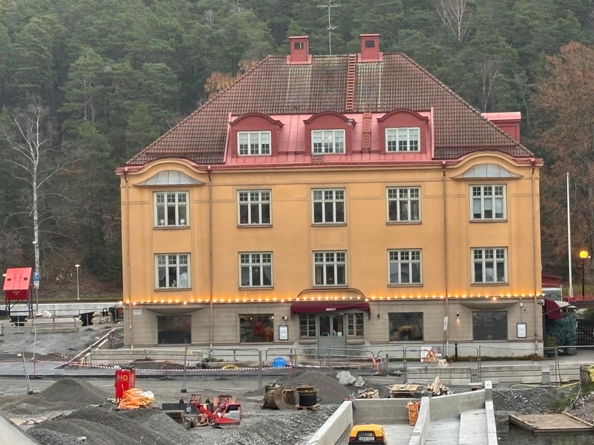 En gul byggnad med rött tak och röda takkupor står i centrum av bilden. Lyser lösa lampor är fästa längs byggnadens fasad på första våningen. På taket syns två skorstenar och en stege. Framför byggnaden pågår ett byggarbete med grushögar, byggnadsred