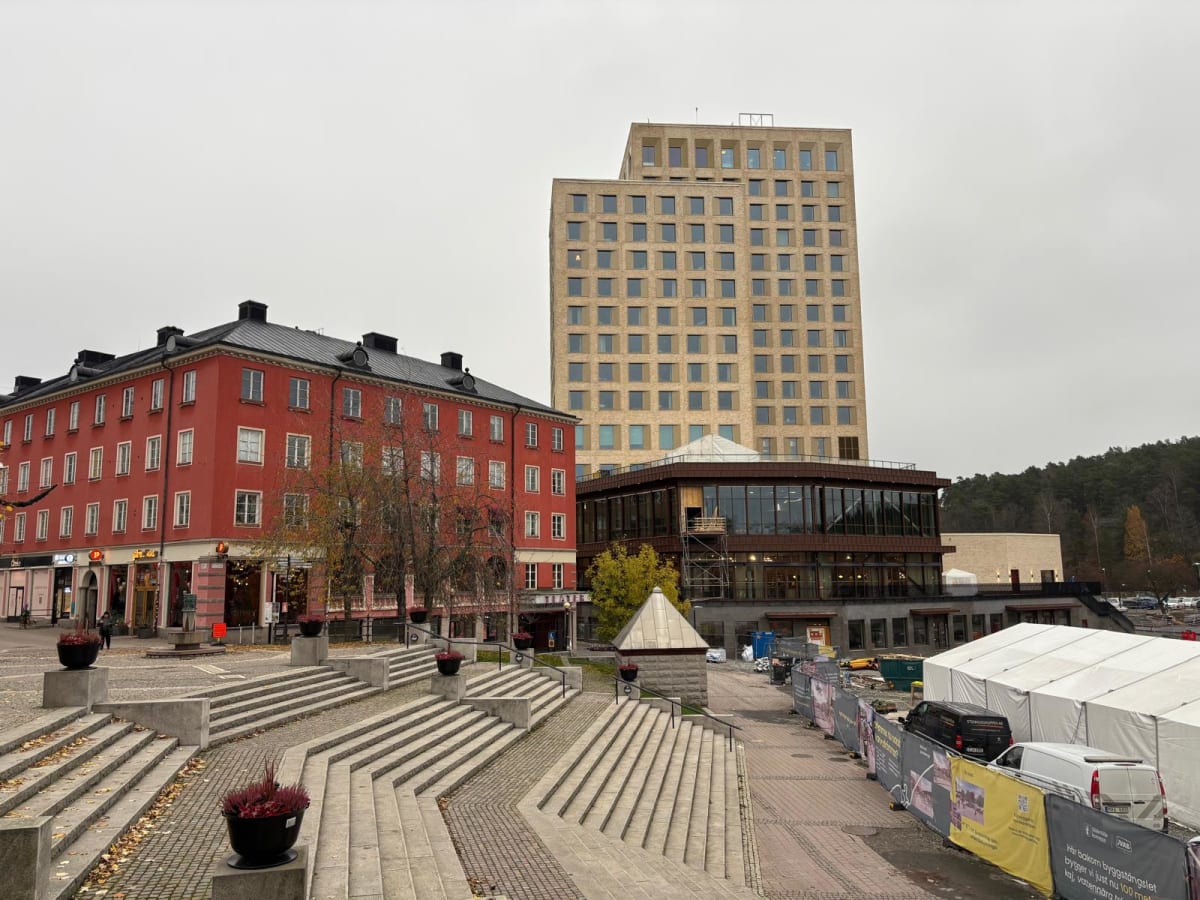Bilden visar ett stadslandskap med flera byggnader under en gråmulen himmel. I bildens centrum till vänster står en röd byggnad med svart tak och vita fönsterkarmar. Framför den röda byggnaden sträcker sig en stor stentrappa med planteringskärl prydd