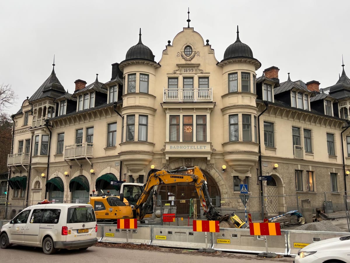 En stor, ljusgul byggnad med texten "BADHOTELLET" och årtalet "1899" syns i bildens centrum. Byggnaden har flera fönster, ett flertal balkonger med vita räcken samt mörka, kupolformade tak. Framför byggnaden pågår ett byggarbete med en gul grävmaskin