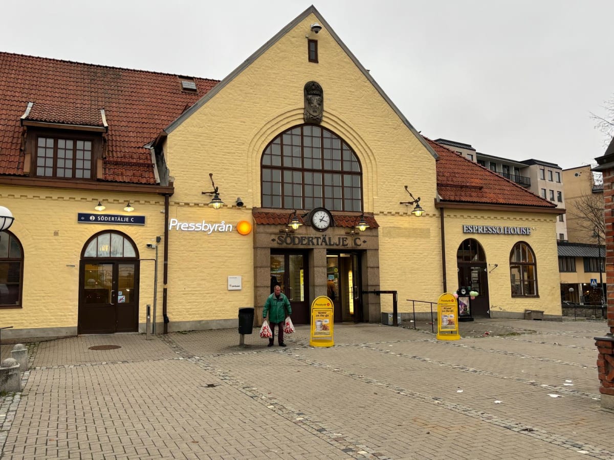 Bilden visar fasaden på en gul byggnad med ett tegeltak under en mulen himmel. Byggnaden har skyltar med texten "SÖDERTÄLJE C", "Pressbyrån" och "Espresso House". En stor, rundad fönstergrupp ses i mitten av den övre delen av byggnaden. Under denna s