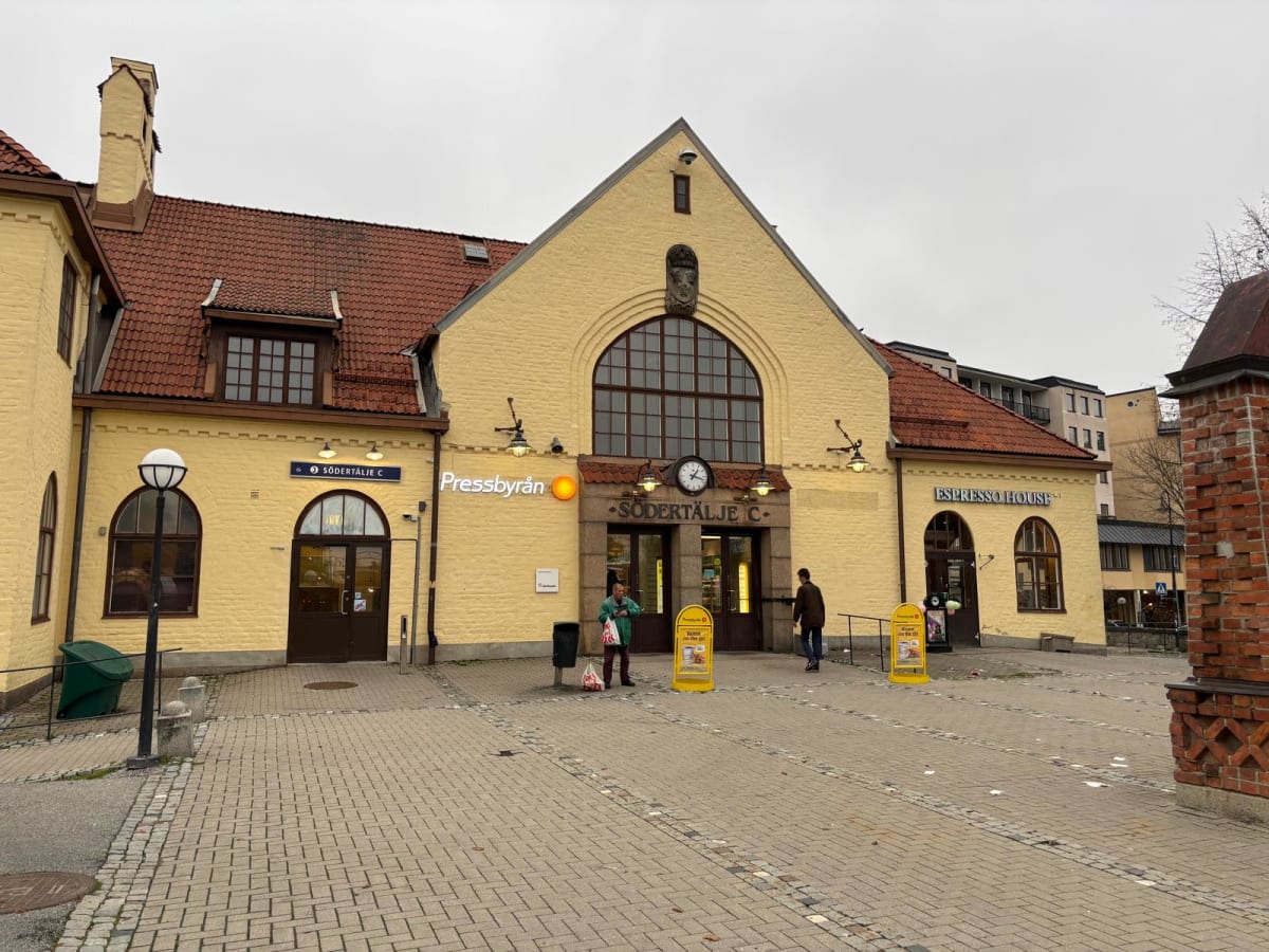 Bilden visar fasaden på en gul tegelbyggnad med röda takpannor. Byggnaden har flera ingångar och fönster i olika storlekar och former. I mitten av byggnaden finns en stor ingång med en digital klocka ovanför. Ovanför ingången står det "Södertälje C"