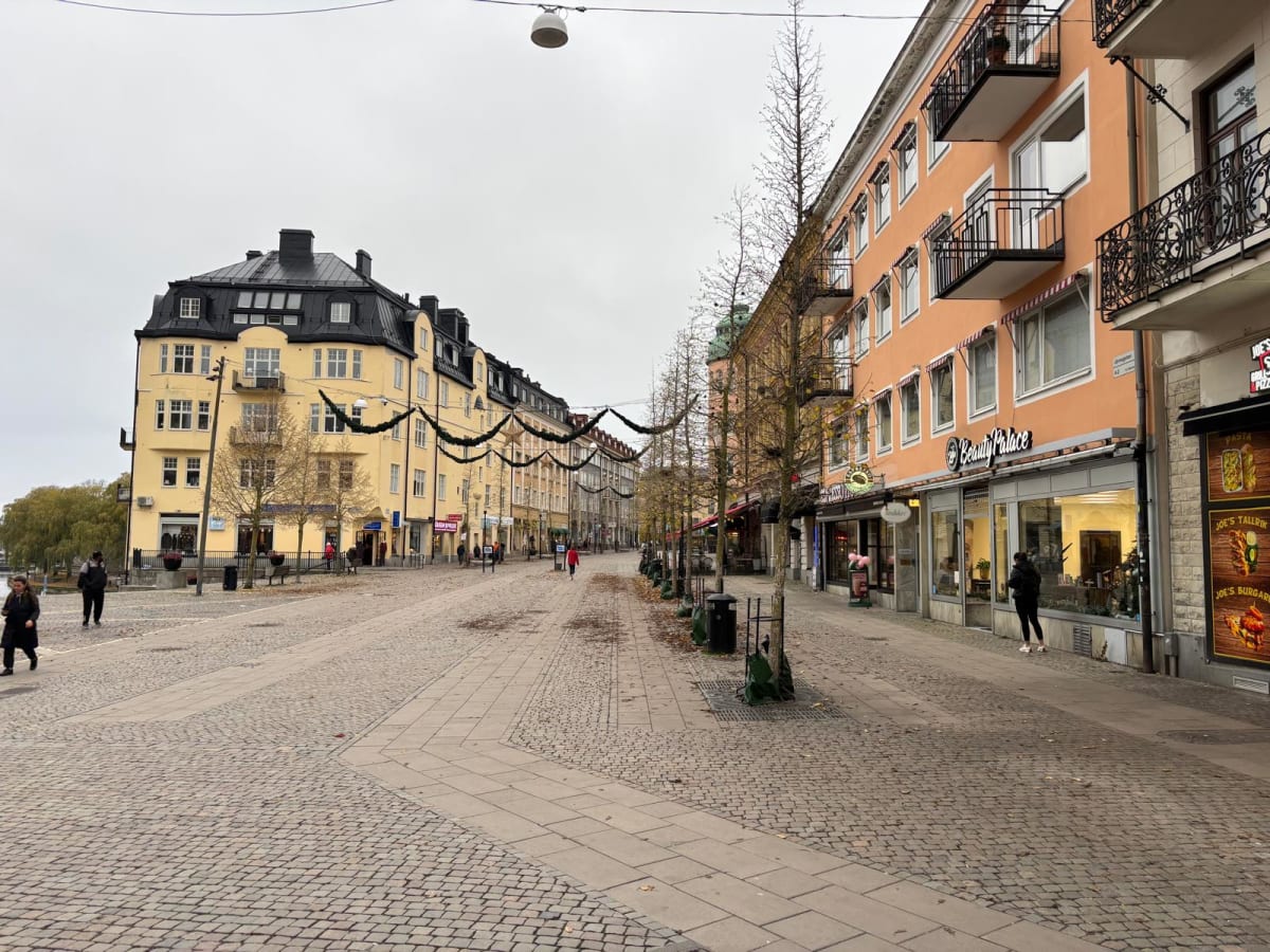 Bilden visar en gata i en stad under dagtid. Gatstensbeläggning täcker större delen av marken, med rektangulära stenplattor som gångvägar. Längs båda sidor av gatan finns flervåningsbyggnader med butiker på bottenvåningen. På den vänstra sidan syns b