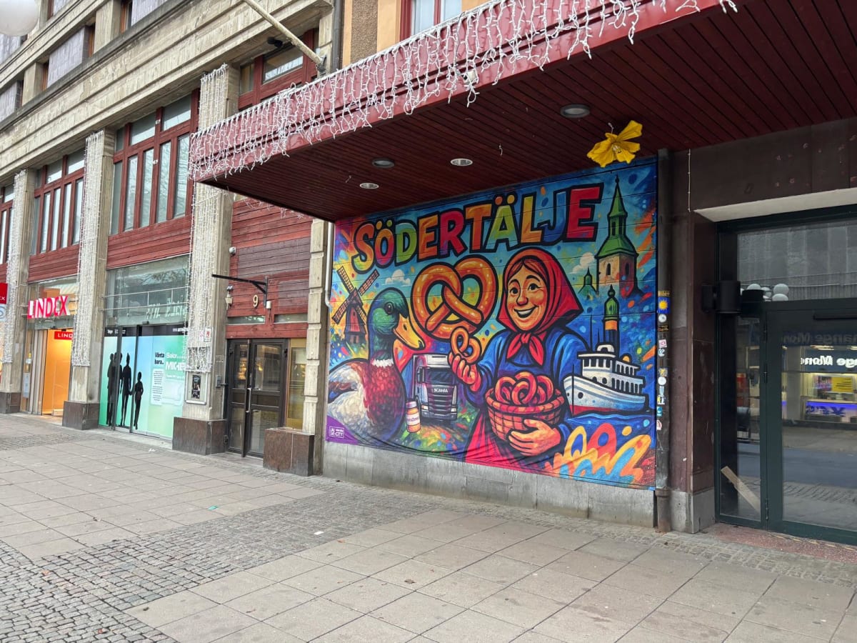 En fasadbild tagen från marknivå visar en färgglad väggmålning som täcker en del av en byggnad längs en stadsgata. Målningen har texten "SÖDERTÄLJE" i stora bokstäver. Den avbildar en kvinna i traditionell klädsel som håller en korg med kringlor, en