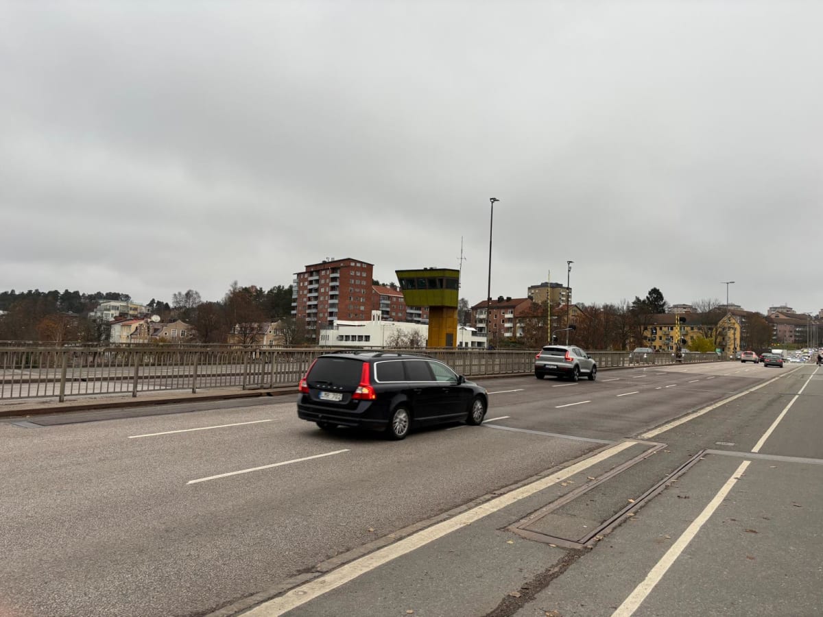 Bilden visar ett gatu- eller broparti med stadsmiljö i bakgrunden. Ett svart personfordon, en kombi, kör bort från betraktaren i vänster fil, medan ett silverfärgat SUV-liknande fordon kör i samma riktning i filen till höger. Ett flertal andra bilar