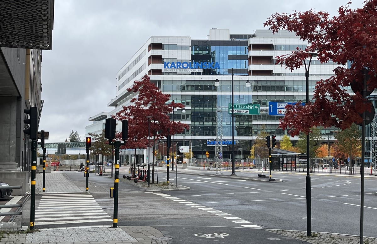 Bilden visar en gata med en byggnad i bakgrunden. Byggnaden har texten "KAROLINSKA UNIVERSITETSSJUKHUSET" på fasaden. På gatan finns trafikljus, en zebraövergång med tydliga vita markeringar, samt vägmarkeringar inklusive en symbol för cykelled. Någr