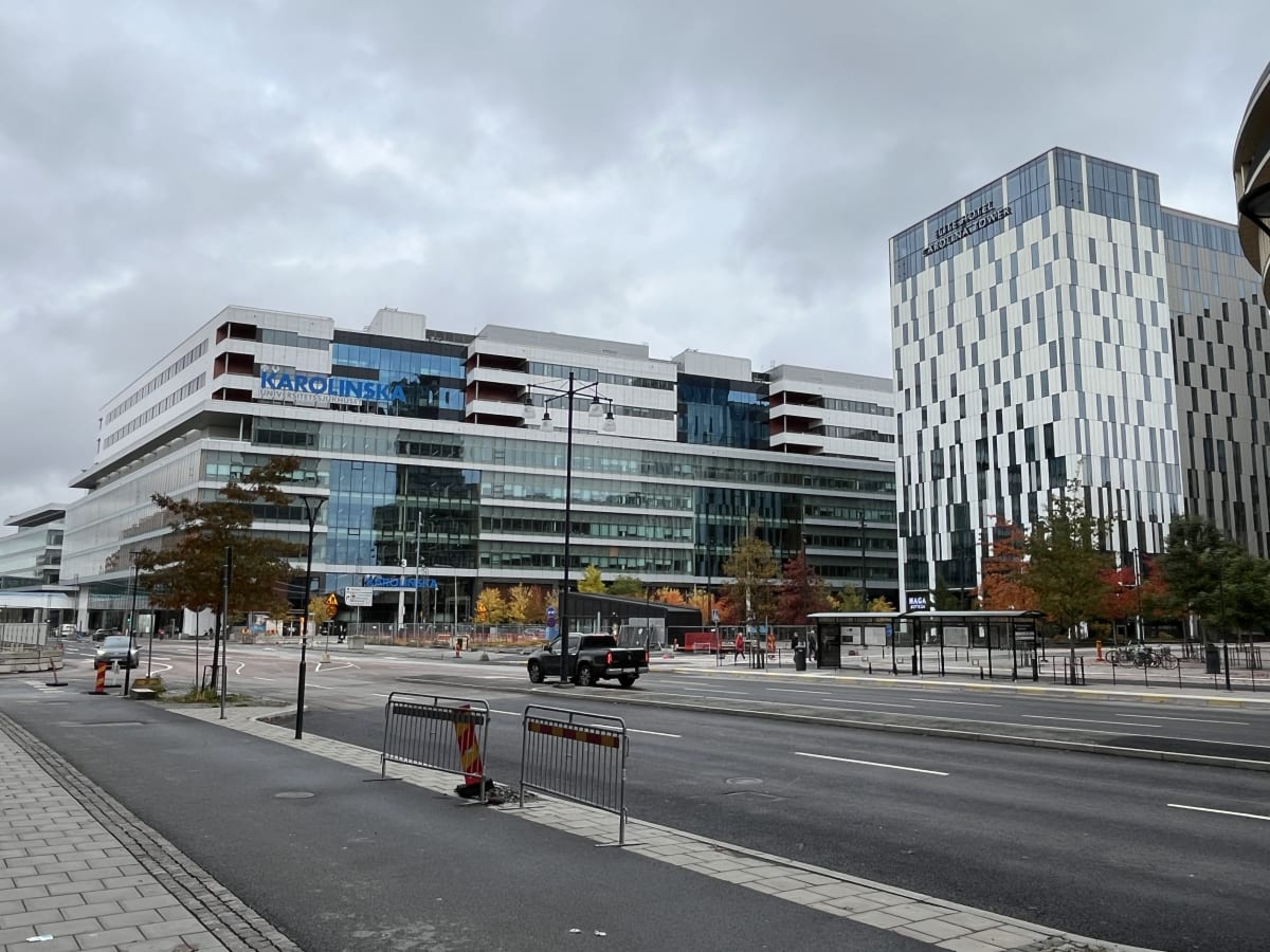 Bilden visar en gata med flera stora byggnader i bakgrunden under en mulen himmel. Till vänster, i mitten av bilden, syns en stor, modern byggnad med texten "KAROLINSKA UNIVERSITETSSJUKHUSET" i blått. Till höger om denna byggnad står en annan hög, mo