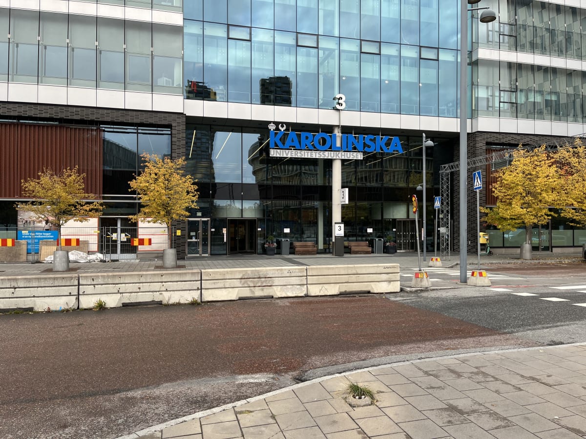 En bild tagen utomhus som visar ingången till en stor glasbyggnad med texten "KAROLINSKA UNIVERSITETSSJUKHUSET" ovanför entrén. Byggnaden har flera våningar med fönster som reflekterar omgivningen. Framför byggnaden finns en trottoar med flera bänkar
